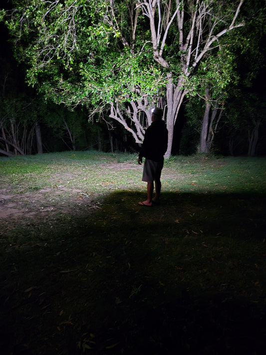 A person standing outdoors at night on a grassy path, photographed in very low light with trees and dark foliage visible on both sides.